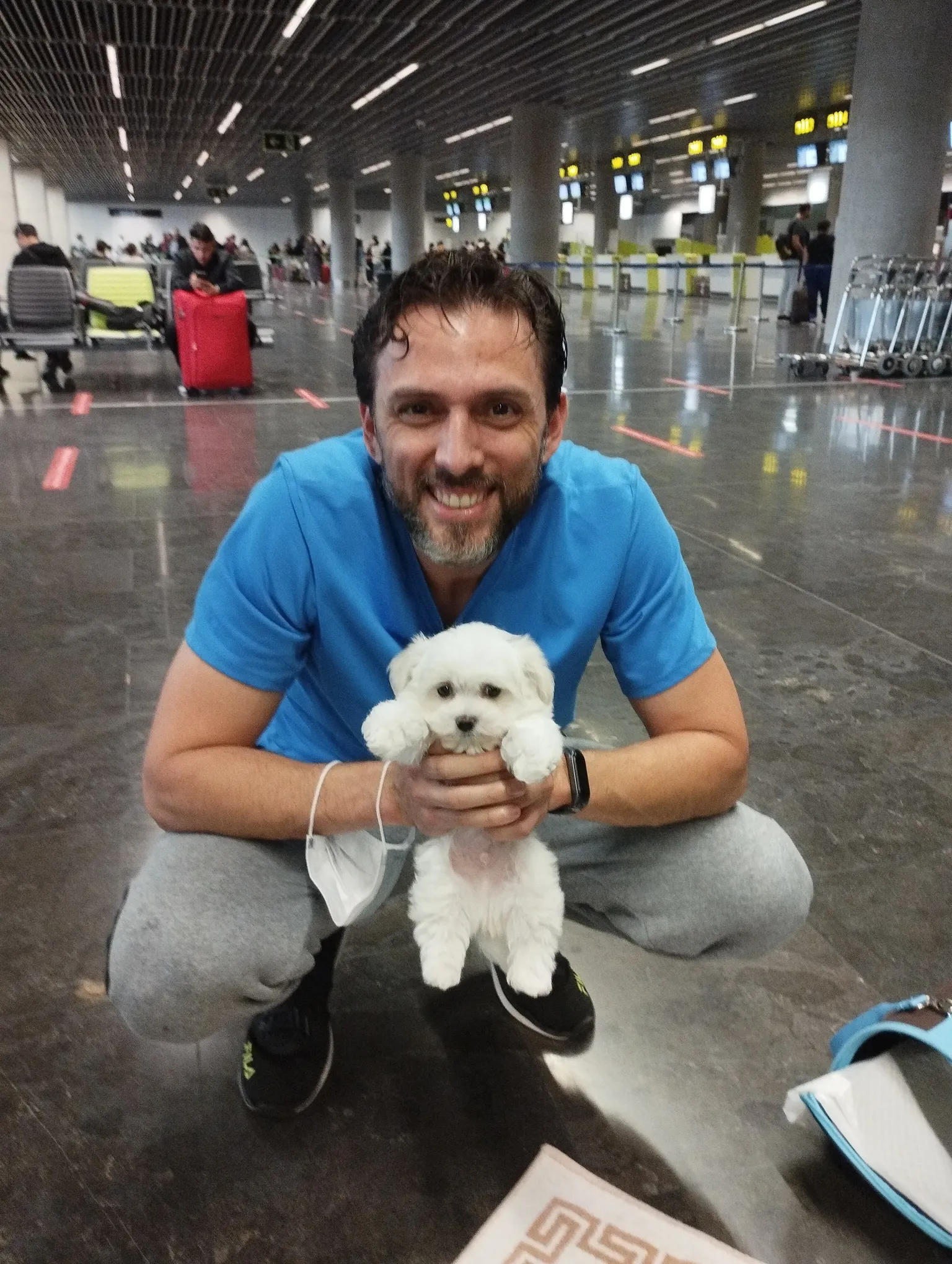 Hombre sonriente en un aeropuerto sosteniendo un cachorro blanco. El fondo muestra una terminal con pasajeros y equipaje, destacando la emoción de la llegada o despedida.