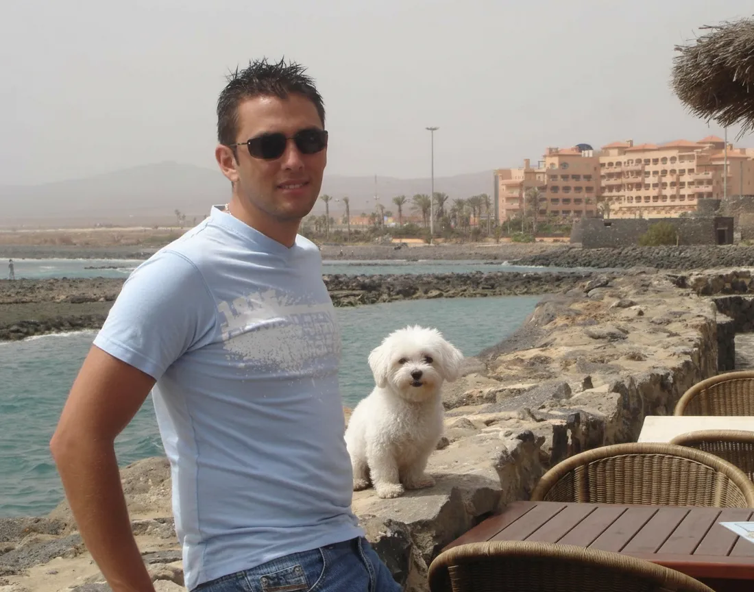 Hombre joven con camiseta azul y gafas de sol posando junto a un perro pequeño en una costa, con vistas al mar y edificios de fondo en un día soleado.