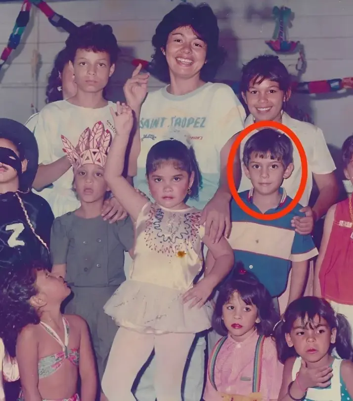 Foto antigua de un grupo de niños y una mujer, todos vestidos con ropa colorida en una celebración. La mujer está en el centro, mientras que los niños muestran diversas expresiones. Un niño con camiseta azul está destacado en un círculo rojo, con otros niños alrededor, algunos en disfraces.